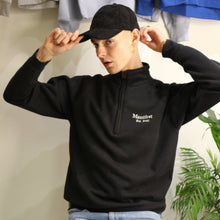 Load image into Gallery viewer, Young man wearing a black Manifest 1/4 zip sweatshirt with embroidered logo, styled with a black cap.