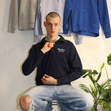 Load image into Gallery viewer, Male model wearing a navy Manifest 1/4 zip sweatshirt in front of hoodies, showcasing embroidered logo.