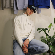 Load image into Gallery viewer, Unisex 1/4 zip sweatshirt in white with Manifest logo, model sitting against a plant and hanging sweatshirts.