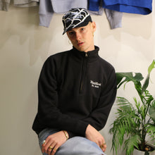 Load image into Gallery viewer, Male model wearing Manifest black 1/4 zip sweatshirt with embroidered logo, styled with a cap and casual accessories.