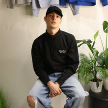 Load image into Gallery viewer, Male model wearing a Manifest black 1/4 zip sweatshirt, featuring embroidered logo, sitting amidst greenery.