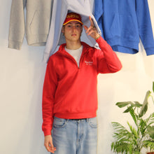 Load image into Gallery viewer, Model wearing a red cotton 1/4 zip sweatshirt with embroidered logo, surrounded by various hoodies in a stylish indoor setting.