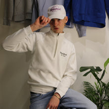 Load image into Gallery viewer, Male model wearing a white Manifest 1/4 zip sweatshirt with embroidered logo, posing in a stylish setting.