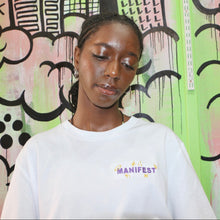 Load image into Gallery viewer, Person wearing a white shirt with 'MANIFEST' logo in front of a colorful graffiti wall.