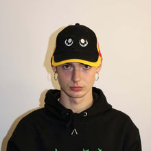 Load image into Gallery viewer, Manifest black cap with bold yellow anime eye embroidery, combining streetwear style and anime inspiration.
