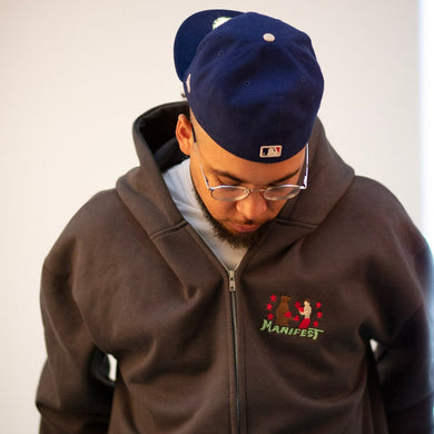 Person wearing a brown hoodie with a logo and text, standing against a plain background