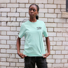 Load image into Gallery viewer, Person wearing a light green t-shirt with text, standing against a white brick wall.
