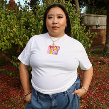 Load image into Gallery viewer, Person wearing a white t-shirt with a graphic design, standing outdoors with greenery and a wooden fence in the background.

