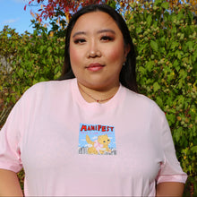 Load image into Gallery viewer, Woman wearing a pink t-shirt with a cartoon cat and 'MANIFEST' text in an outdoor setting.
